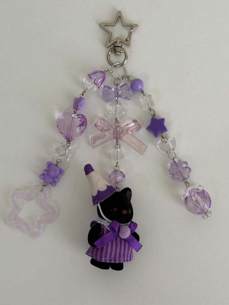 May include: A keychain featuring a star-shaped metal clasp and three dangling strands of purple and clear beads, including heart, star, and bear shapes, plus bows. A small black animal figure wearing a purple striped dress and party hat hangs below.