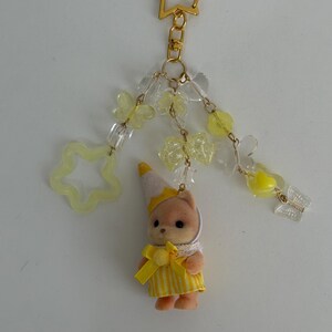 May include: A keychain featuring a small flocked animal figurine wearing a yellow striped dress and a party hat. The charm is attached to a gold star-shaped clasp with dangling yellow and clear beads in butterfly, star, and bow shapes.