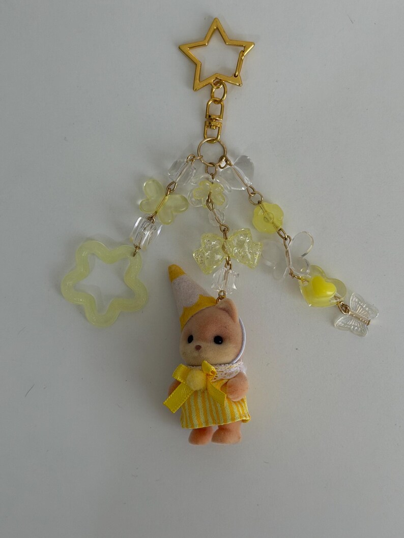 May include: A keychain featuring a small flocked animal figurine wearing a yellow striped dress and a party hat. The charm is attached to a gold star-shaped clasp with dangling yellow and clear beads in butterfly, star, and bow shapes.