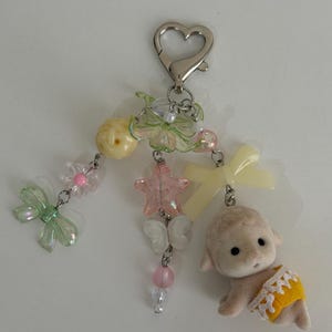 May include: A keychain featuring a silver heart-shaped clasp attached to three dangling strands of beads. The beads include light green bows, a yellow sphere, a pink star, white butterflies, and a small flocked lamb figure wearing a yellow diaper.