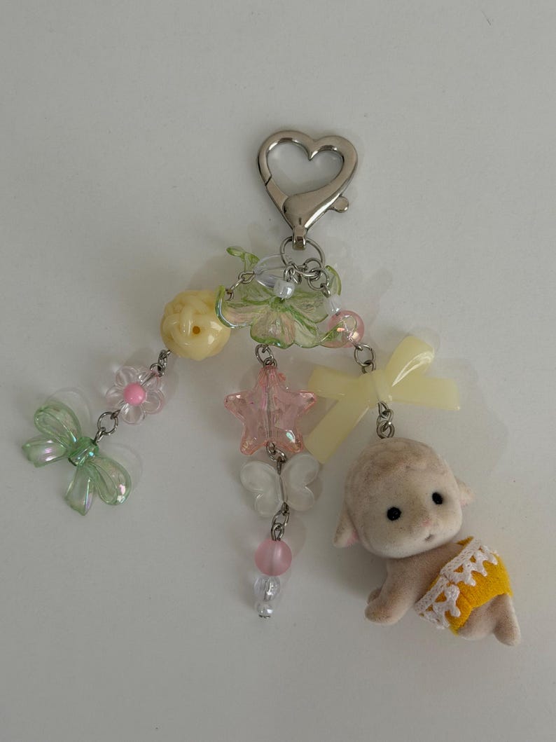 May include: A keychain featuring a silver heart-shaped clasp attached to three dangling strands of beads. The beads include light green bows, a yellow sphere, a pink star, white butterflies, and a small flocked lamb figure wearing a yellow diaper.