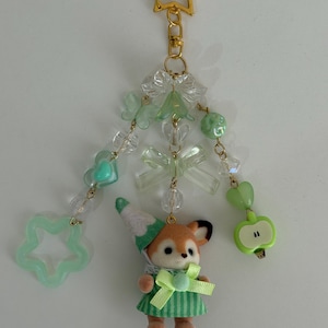 May include: A decorative keychain featuring a gold star clasp, three dangling beaded strands with light green and clear acrylic beads, butterfly and bow charms, a light green apple charm, and a small flocked animal figure wearing a green striped dress.