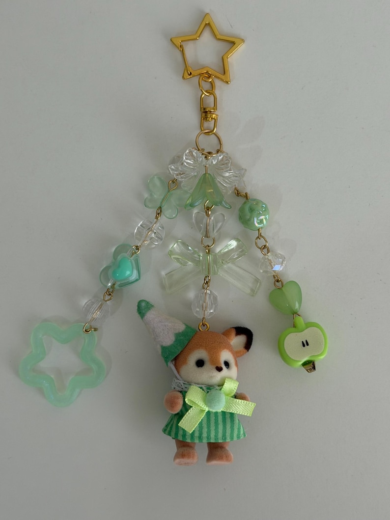 May include: A decorative keychain featuring a gold star clasp, three dangling beaded strands with light green and clear acrylic beads, butterfly and bow charms, a light green apple charm, and a small flocked animal figure wearing a green striped dress.