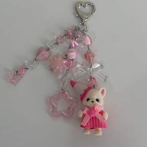 May include: A keychain featuring a silver heart-shaped clasp with multiple dangling strands of pink and clear beads, including heart, star, and butterfly shapes, a clear bow, and a small animal figurine wearing a pink striped dress and hat.