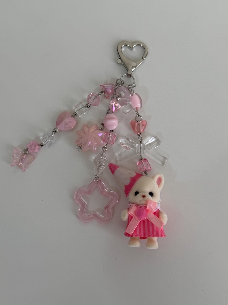 May include: A keychain featuring a silver heart-shaped clasp with multiple dangling strands of pink and clear beads, including heart, star, and butterfly shapes, a clear bow, and a small animal figurine wearing a pink striped dress and hat.