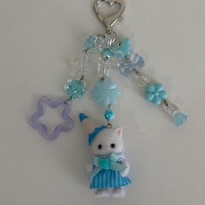 May include: A keychain featuring a silver heart-shaped clasp, light blue and clear beads in flower and star shapes, and a small white cat figurine wearing a blue striped dress and a matching blue party hat with a light blue ribbon bow.