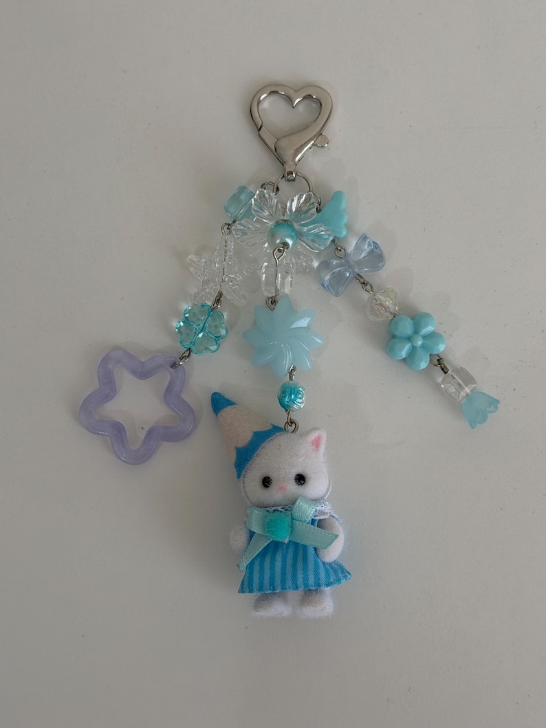 May include: A keychain featuring a silver heart-shaped clasp, light blue and clear beads in flower and star shapes, and a small white cat figurine wearing a blue striped dress and a matching blue party hat with a light blue ribbon bow.