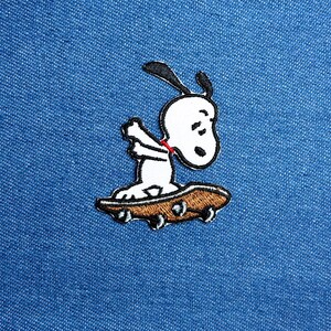 May include: Embroidered patch featuring Snoopy on a skateboard. The patch has a white Snoopy with black ears and a red collar, riding a brown skateboard with black wheels. The patch is on a blue denim background.