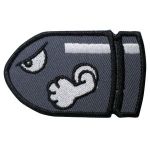 May include: A gray embroidered patch of a bullet bill from the Super Mario Bros. video game series. The bullet bill has a white eye and a white arm.