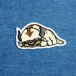 Könnte beinhalten: Gestickter Aufnäher mit einem schlafenden Cartoon-Bison-Charakter. Der Aufnäher hat einen weißen Rand und ist vor einem blauen Jeans-Hintergrund platziert. Das Bison ist hauptsächlich beige mit braunen und grauen Akzenten.