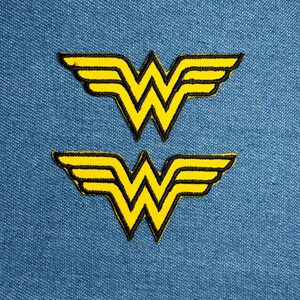 Può includere: Due toppe ricamate con il logo di Wonder Woman. Il logo è una "W" gialla con ali, bordata di nero. Le toppe sono su uno sfondo di denim blu.