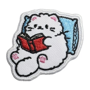 Op de afbeelding: Witte geborduurde patch van een kat die in bed een rood boek leest. De kat ligt op zijn zij met zijn hoofd op een blauw kussen.