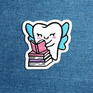 Könnte beinhalten: Gestickter Aufnäher mit einem lächelnden Zahn mit blauen Feenflügeln, der ein rosa Buch auf einem Stapel von Büchern liest. Der Aufnäher hat einen weißen Rand, vor blauem Jeansstoff.