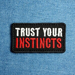 May include: A rectangular black embroidered patch with the words "TRUST YOUR INSTINCTS" in white and red thread. The patch is set against a blue denim background.