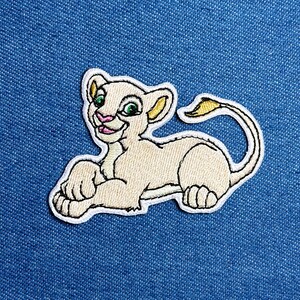 May include: Embroidered patch of a light tan lion cub with green eyes, pink nose, and a yellow-tipped tail. The lion cub is lying down with its paws forward. The patch has a white border and is on a blue denim background.