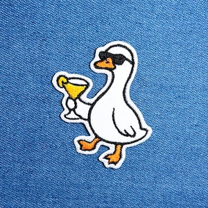 Puede incluir: Parche bordado con un ganso blanco con gafas de sol y un cóctel amarillo. El ganso tiene patas y pico naranjas. El parche está delineado en blanco y negro, sobre un fondo de mezclilla azul.