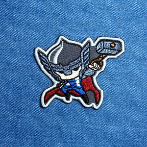 Peut inclure: Écusson brodé représentant Thor, le super-héros de Marvel Comics, dans un style cartoon. L'écusson montre Thor avec son casque, sa cape et son marteau. L'écusson est bordé de blanc et posé sur un fond en denim bleu.