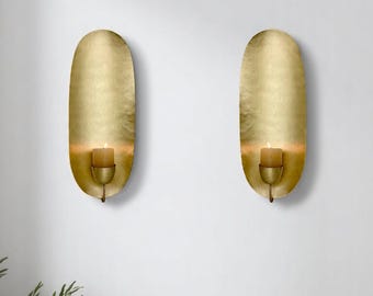 May include: A set of two gold metal wall sconces with a candle holder in the center of each. The sconces are oval shaped and have a hammered metal finish.