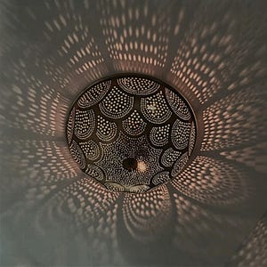 May include: A decorative, round, bronze-colored pendant light fixture with a detailed, dotted pattern. The light casts intricate shadows on the surrounding surface, creating a play of light and shadow. The fixture is designed to hang from the ceiling.