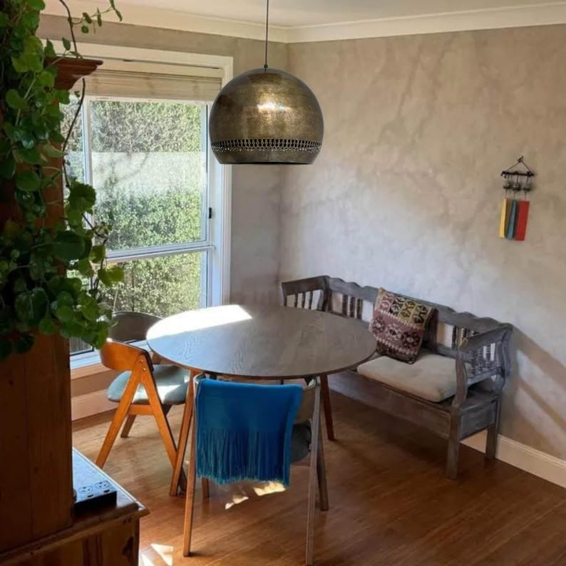 Op de afbeelding: Een ronde, metalen, goudkleurige hanglamp hangt boven een houten tafel met stoelen in een eetkamer. De muren zijn lichtgrijs geverfd.