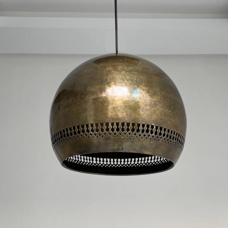 Op de afbeelding: Een ronde, messing hanglamp met een gehamerde afwerking en een decoratief uitsparingontwerp rond de onderrand.
