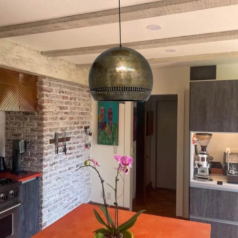 Op de afbeelding: Een ronde, goudkleurige hanglamp met een decoratief patroon rond de onderrand hangt aan een zwart snoer in een keuken.