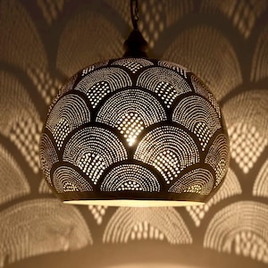 Pode incluir: Uma luminária pendente de metal em forma de cúpula com um padrão perfurado de semicírculos concêntricos. A superfície externa escura é perfurada com numerosos pequenos orifícios que deixam passar a luz, projetando uma sombra de arco.