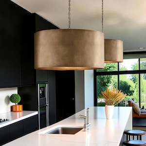 May include: Two large, cylindrical pendant lights with a brushed bronze finish hang from thin metal chains. The drum-shaped shades feature a smooth, metallic surface that reflects light, suspended above a white kitchen island countertop.