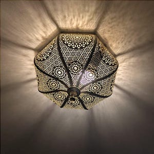 May include: A decorative ceiling light fixture with a geometric, intricate design. The light is made of metal and has a warm, golden finish. The light is on and casting a warm glow.