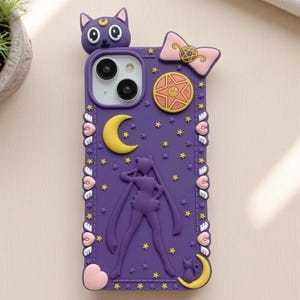 Può includere: Custodia per telefono viola con un design 3D che presenta Sailor Moon, Luna, una luna crescente, stelle e cuori. La custodia ha un fiocco rosa e un ciondolo a forma di stella dorata.