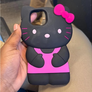 May include: Black phone case featuring a cartoon cat design with a pink bow, dress, and facial details. The case has a cutout for the camera and is held in a hand.