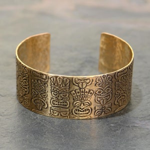 May include: A gold-toned metal bracelet with a repeating pattern of tiki faces and designs.