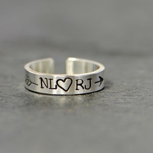 May include: Silver ring with an arrow pointing to the right, a heart, and the initials "NL" and "RJ" engraved on the band.