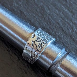 Może przedstawiać: Srebrny pierścień z detalicznym wzorem gór i drzew. Pierścień znajduje się na srebrnym pręcie.