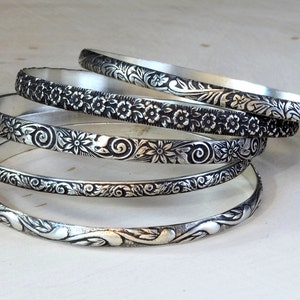 May include: Five silver bangles with intricate floral designs. The bangles are stacked on top of each other, with the most ornate design on the bottom.