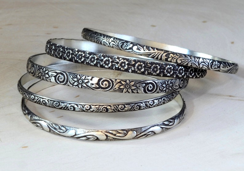May include: Five silver bangles with intricate floral designs. The bangles are stacked on top of each other, with the most ornate design on the bottom.