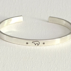 May include: A silver-colored cuff bracelet with a minimalist design. The bracelet features a small bear silhouette and two asterisks engraved on the front. The open-ended design allows for easy wear.