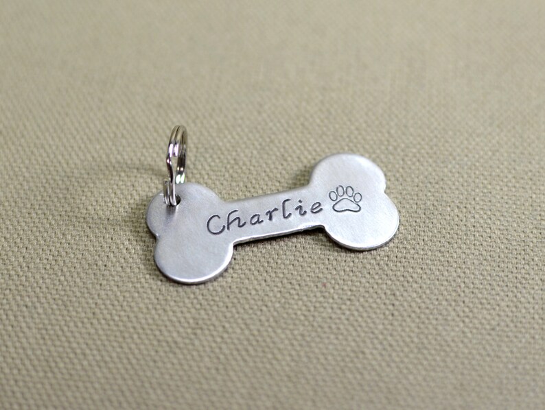 May include: Silver bone-shaped dog tag with a paw print and the name "Charlie" engraved on it.