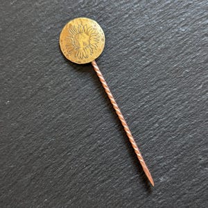 May include: A vintage hat pin with a sunflower design. The pin has a round, gold-coloured head with an etched sunflower and a twisted copper-coloured stem. The pin is set against a dark, textured background.