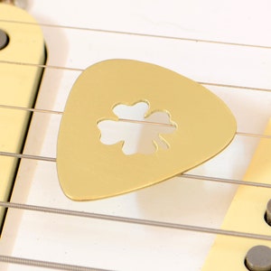 May include: A gold-colored guitar pick with a four-leaf clover cut-out design. The pick is resting on the strings of a guitar.