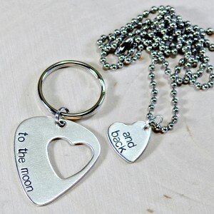 Op de afbeelding: Een zilverkleurige sleutelhanger met een plectrumvorm en een hartuitsparing, gegraveerd met "to the moon". Een bijpassende hartvormige hanger met "and back" aan een kogelketting.