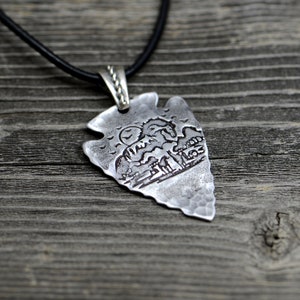 Puede incluir: Un collar con un colgante de punta de flecha de plata, con un diseño grabado detallado de una escena de montaña con un sol y pájaros. El collar está en un cordón de cuero negro.