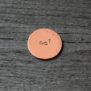 May include: A copper-colored round disc with a hammered texture and the infinity symbol followed by the number "7" engraved on the surface.