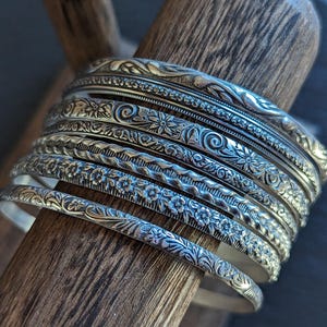 May include: A set of nine silver bangles with intricate floral and geometric designs. The bangles are stacked on top of each other, creating a layered look.