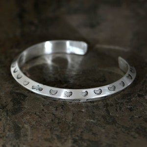 May include: A silver bracelet featuring a decorative pattern of small, repeating shapes. The bracelet is open and has a smooth, polished finish.