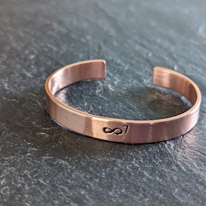 Op de afbeelding: Een koperen manchetarmband met een open ontwerp. De armband heeft een geborstelde afwerking en is voorzien van het nummer "87" dat aan de voorkant is gestempeld. De armband is ongeveer 1,3 cm breed.