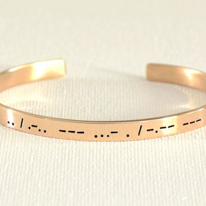 May include: A rose gold bangle bracelet with a message engraved in braille. The message is "I love you".