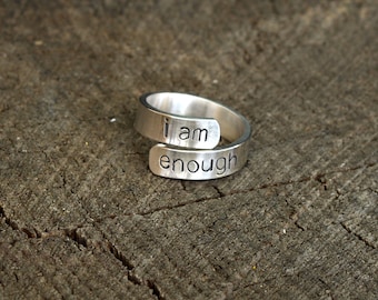 May include: A silver ring with the words "i am enough" engraved on it. The ring is open and can be adjusted to fit different finger sizes.