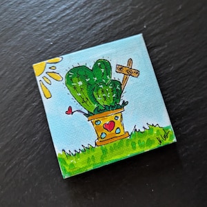 Op de afbeelding: Een kleine vierkante canvas schildering met een cartoon cactus in een gele pot met een hart. De cactus heeft hartvormige segmenten en staat op groen gras onder een gele zon. Een klein houten bord staat naast de cactus.