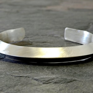 May include: A brushed silver cuff bracelet. The open-ended bracelet has a wide, flat band that tapers slightly towards the ends. The metal has a matte finish, reflecting light subtly. The bracelet is designed to be worn on the wrist.